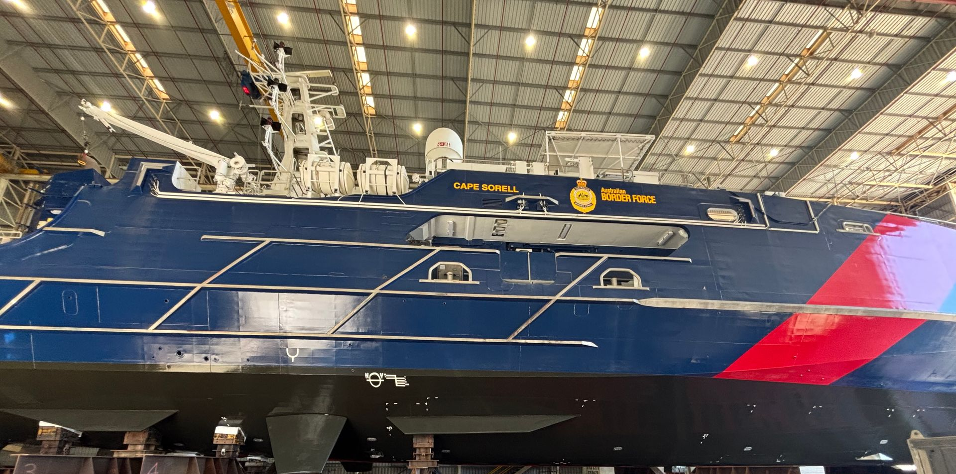 Blue racing sailboat on display indoors, with red bow and white mast under a hangar ceiling
