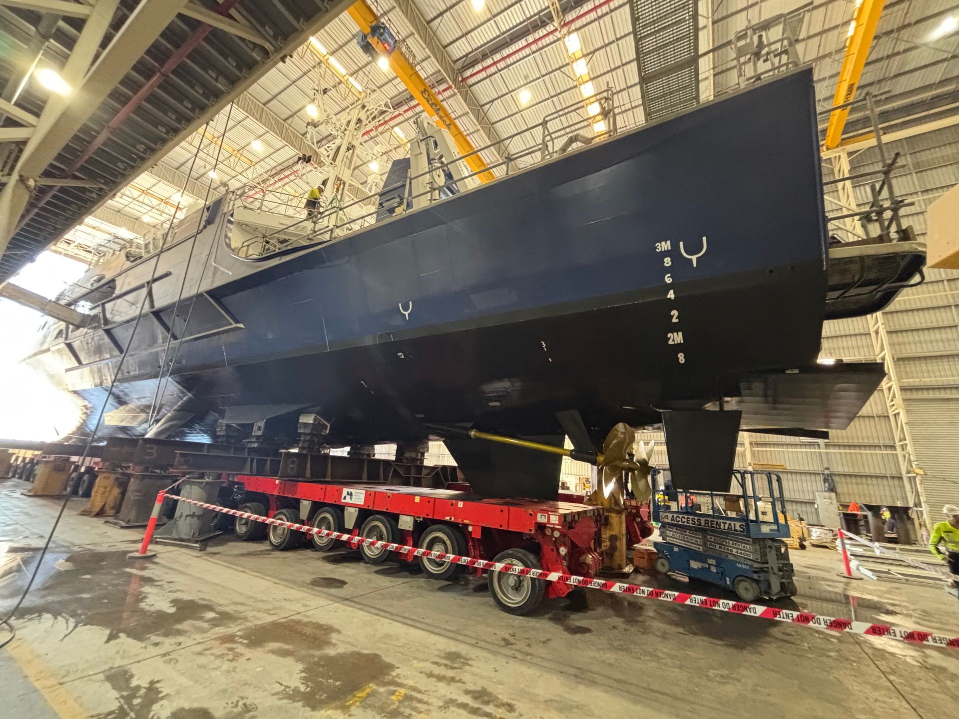 Large black boat hull on a red trailer inside an industrial warehouse, under bright overhead lights