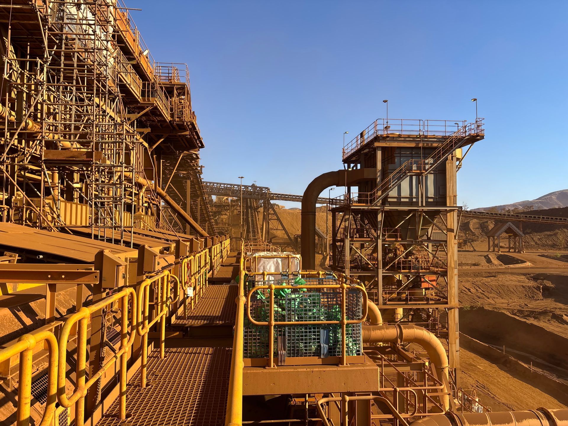 Industrial mining facility with rust-colored metal structures, pipes, and conveyors in a dusty open-pit site