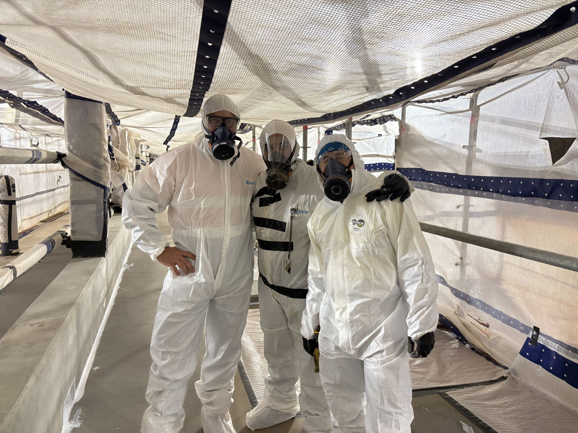 Three people in white protective suits walking through a tunnel-like corridor.