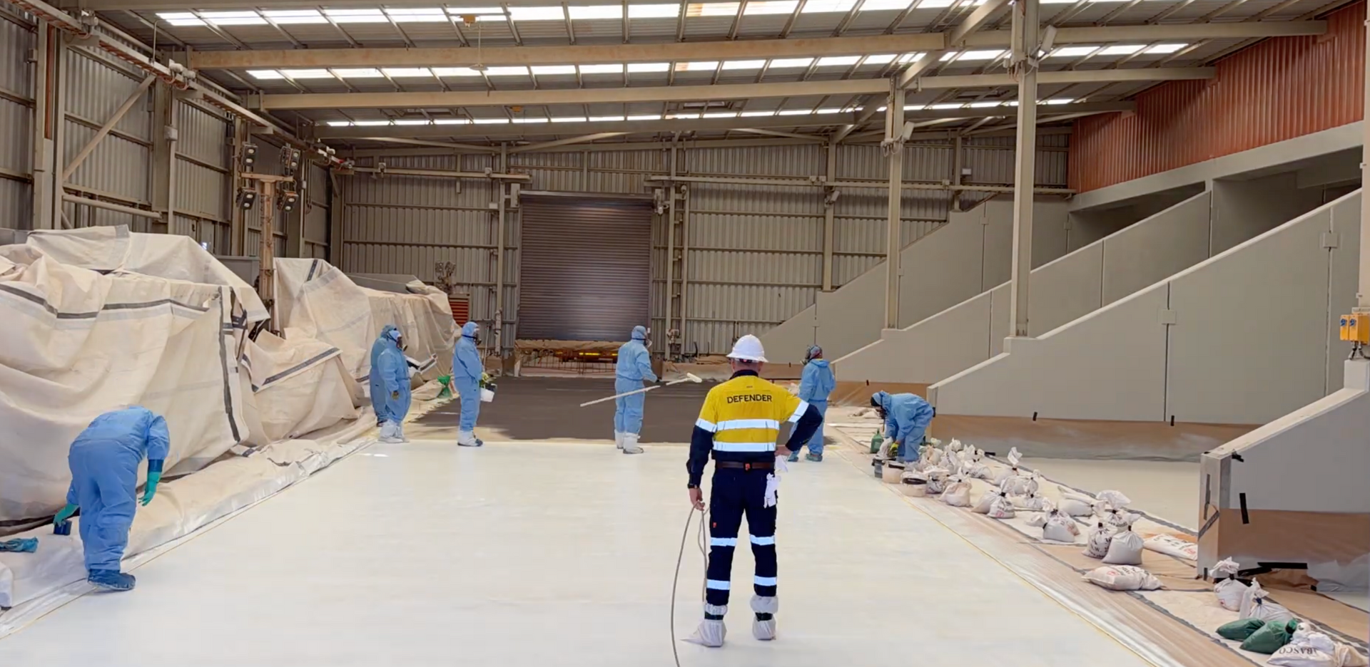 Workers in blue coveralls spray-coating a large industrial hall floor and walls