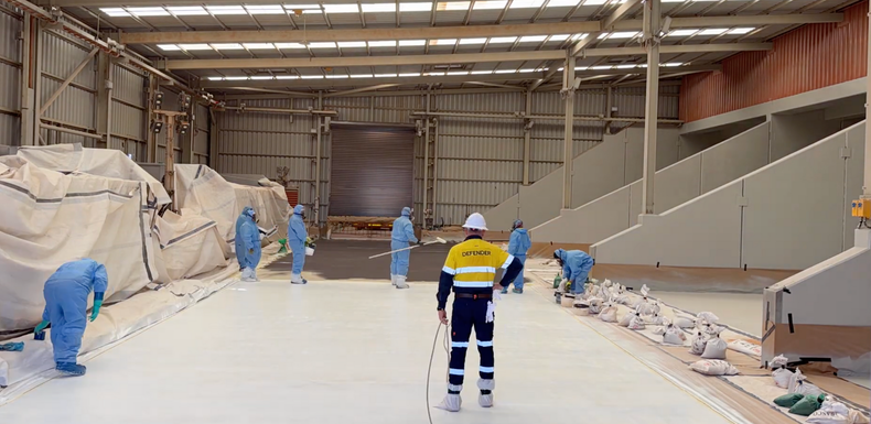 Workers in blue coveralls spray-coating a large industrial hall floor and walls