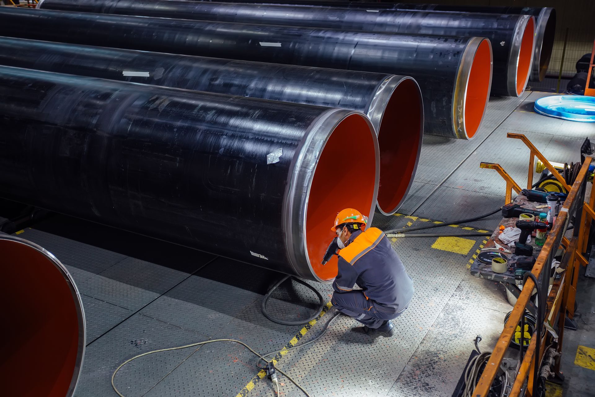 Worker welds a large black pipeline section in an industrial facility, with bright orange pipe ends.