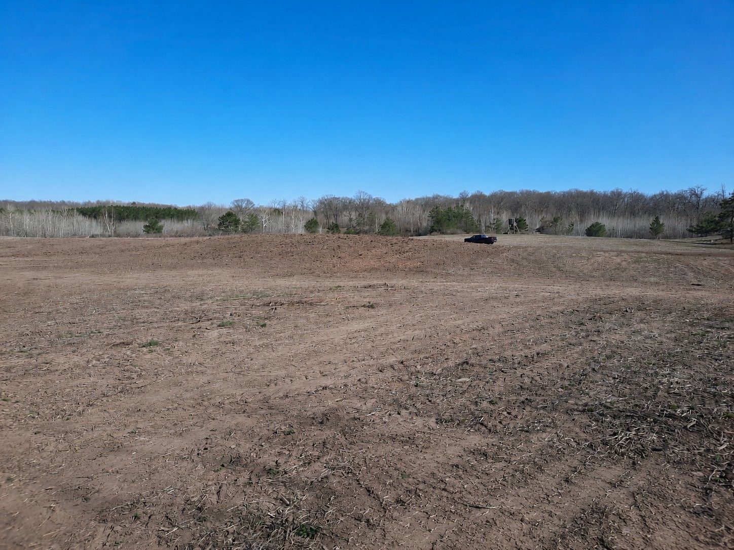 Dry, plowed field under a bright blue sky with a distant tree line and a small dark vehicle near the horizon