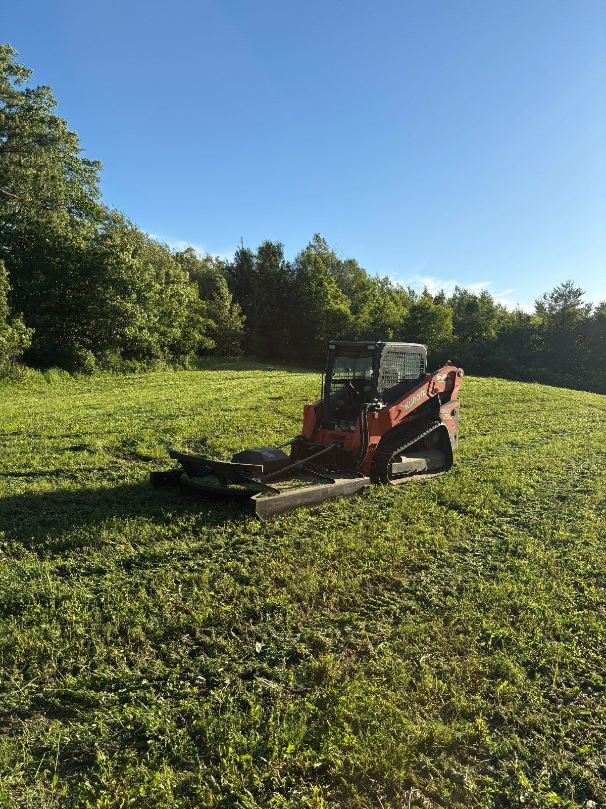 Red skid steer mower clearing a grassy field under a clear blue sky
