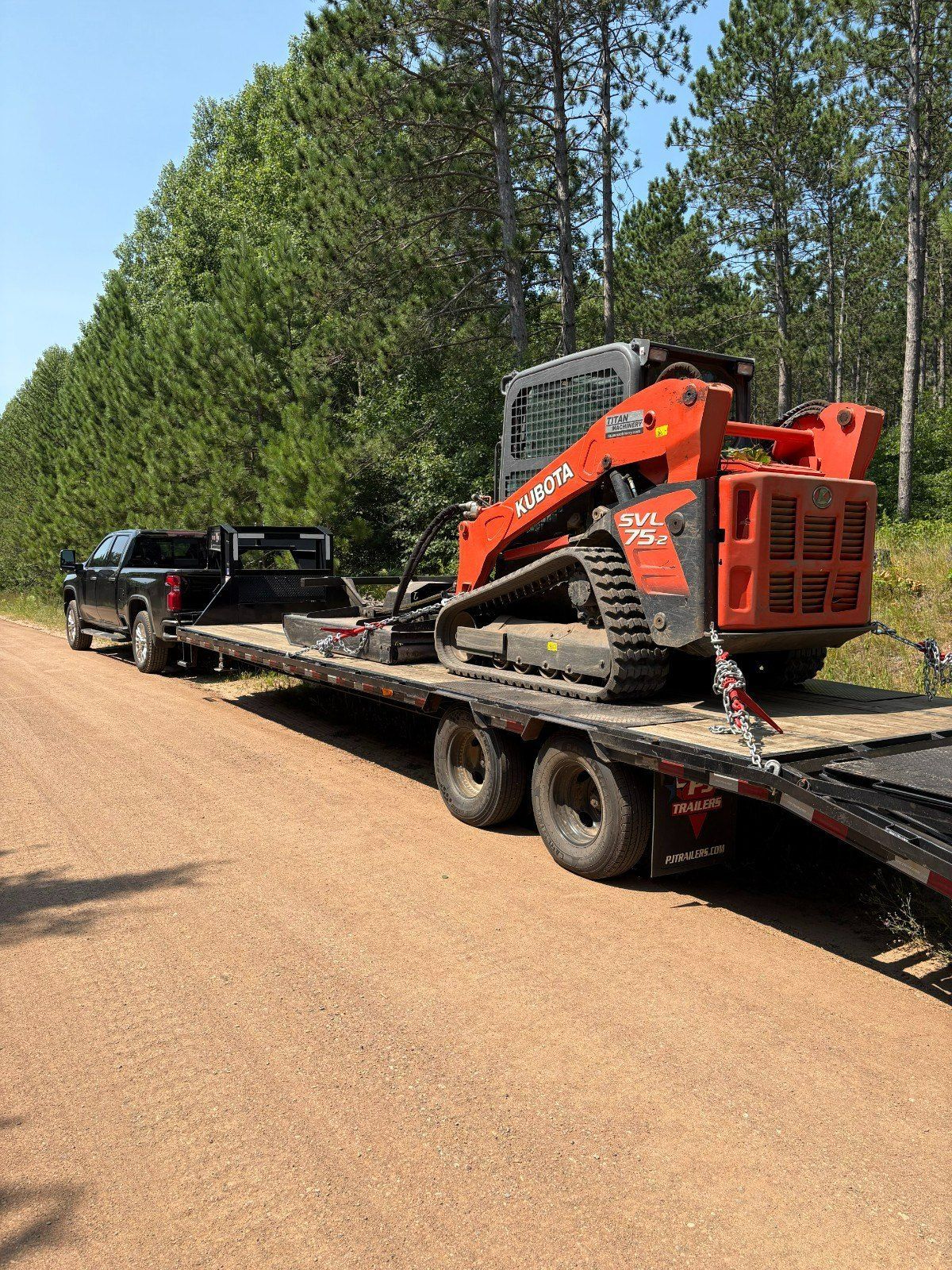 Orange skid steer on a flatbed trailer behind a black pickup on a gravel road, trees in the background