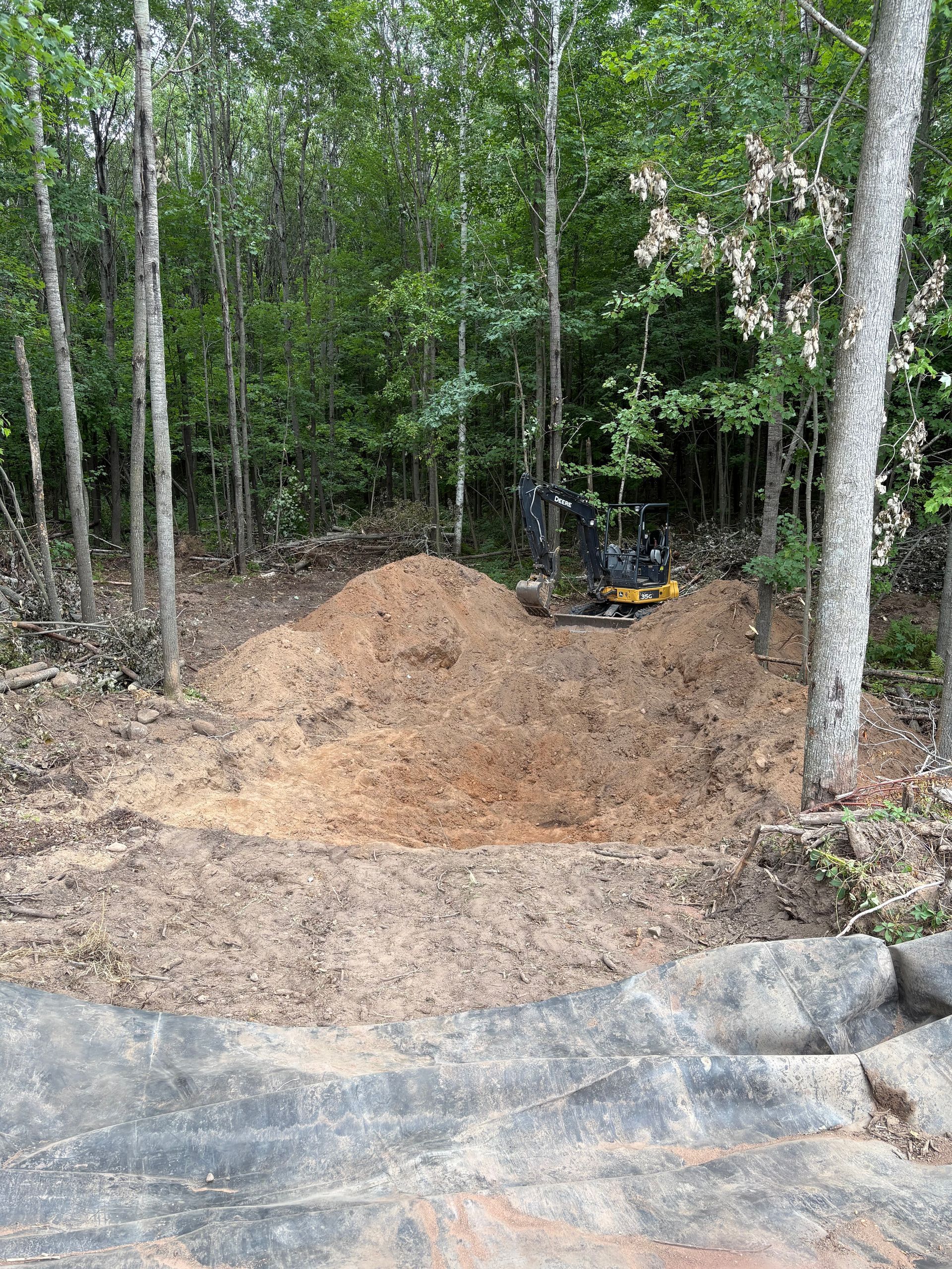 Fresh dirt mound beside a small excavator in a wooded clearing with large rocks in the foreground