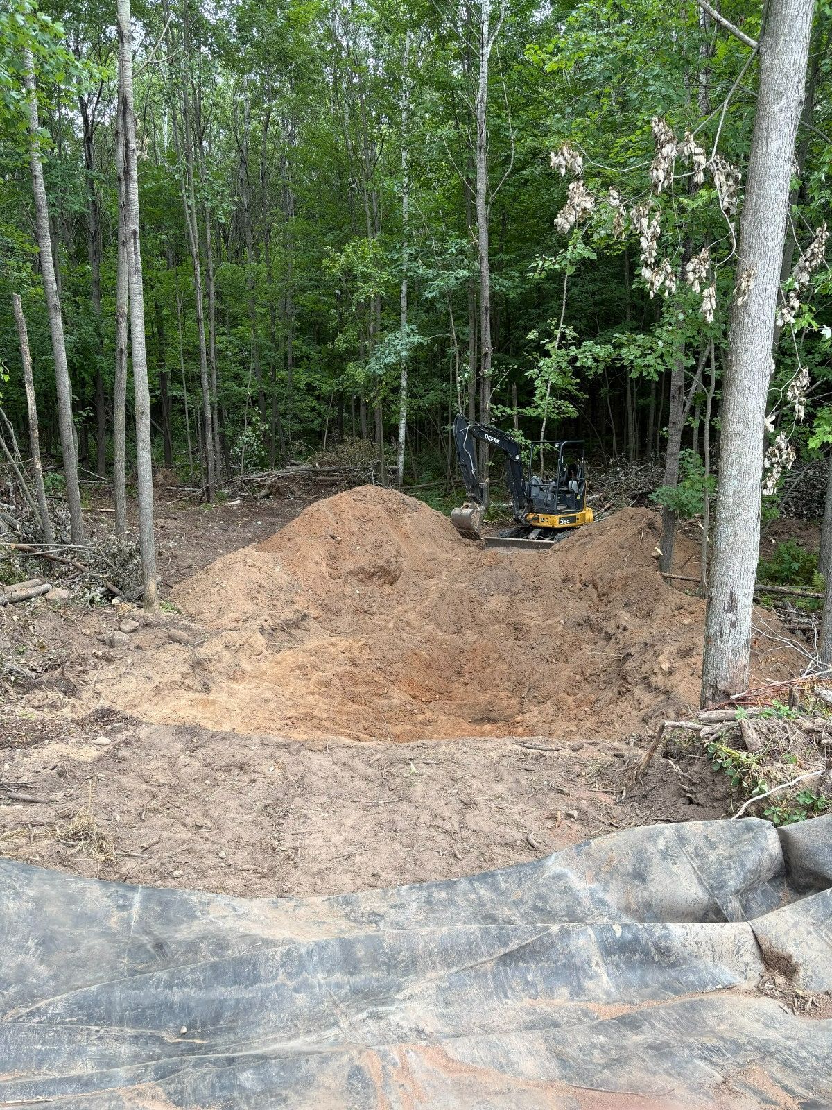 Mound of fresh dirt beside a wooded trail with a small excavator in the background