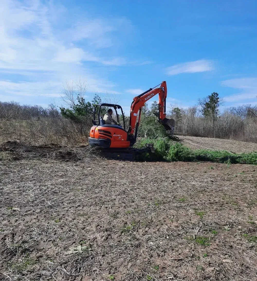 Orange excavator clearing brush at the edge of a field under a blue sky