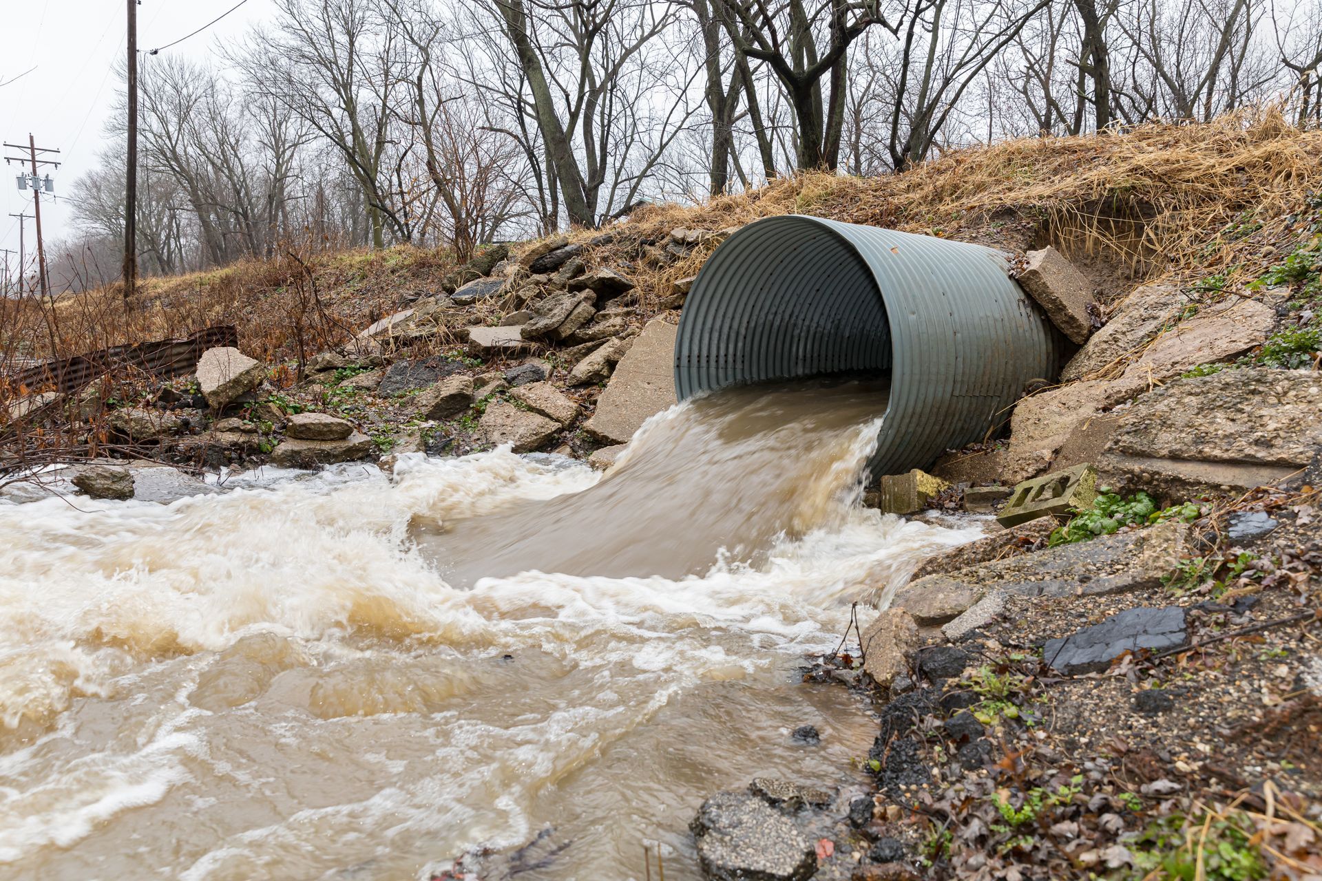 Water rushing from a corrugated metal culvert into a muddy creek beside a rocky embankment