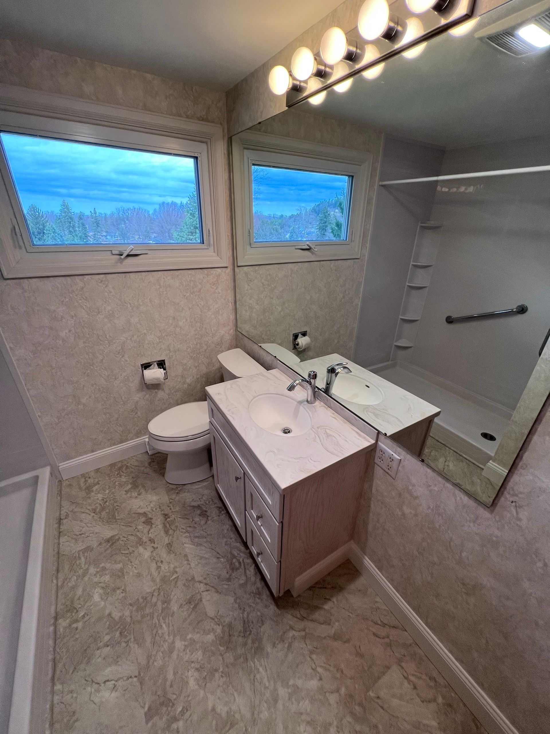Small bathroom with a sink vanity, toilet, mirror, shower, and two windows at dusk.