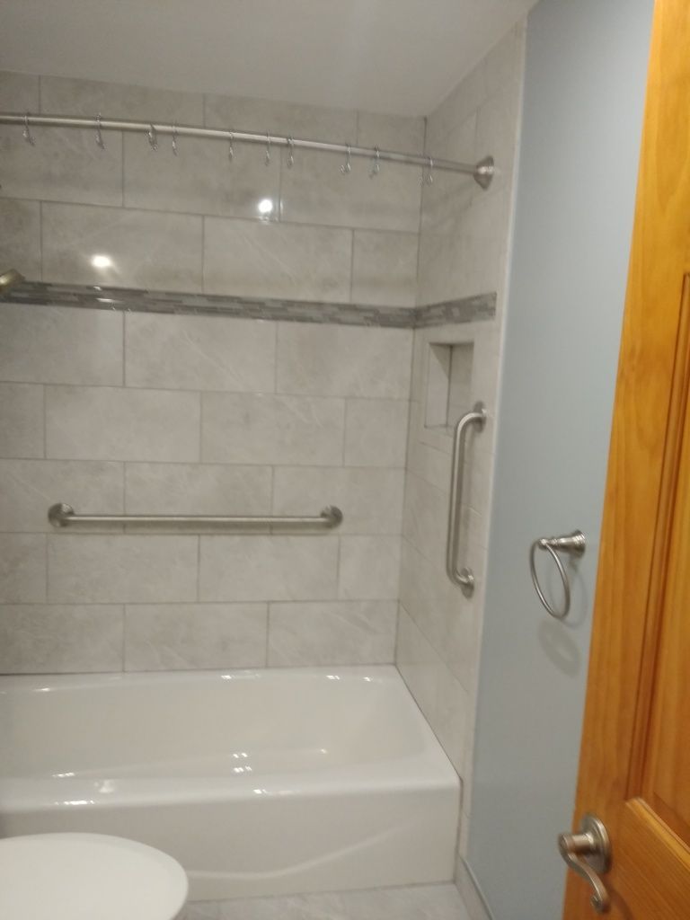 Small bathroom with a white tub, tiled shower walls, grab bars, and a glass door