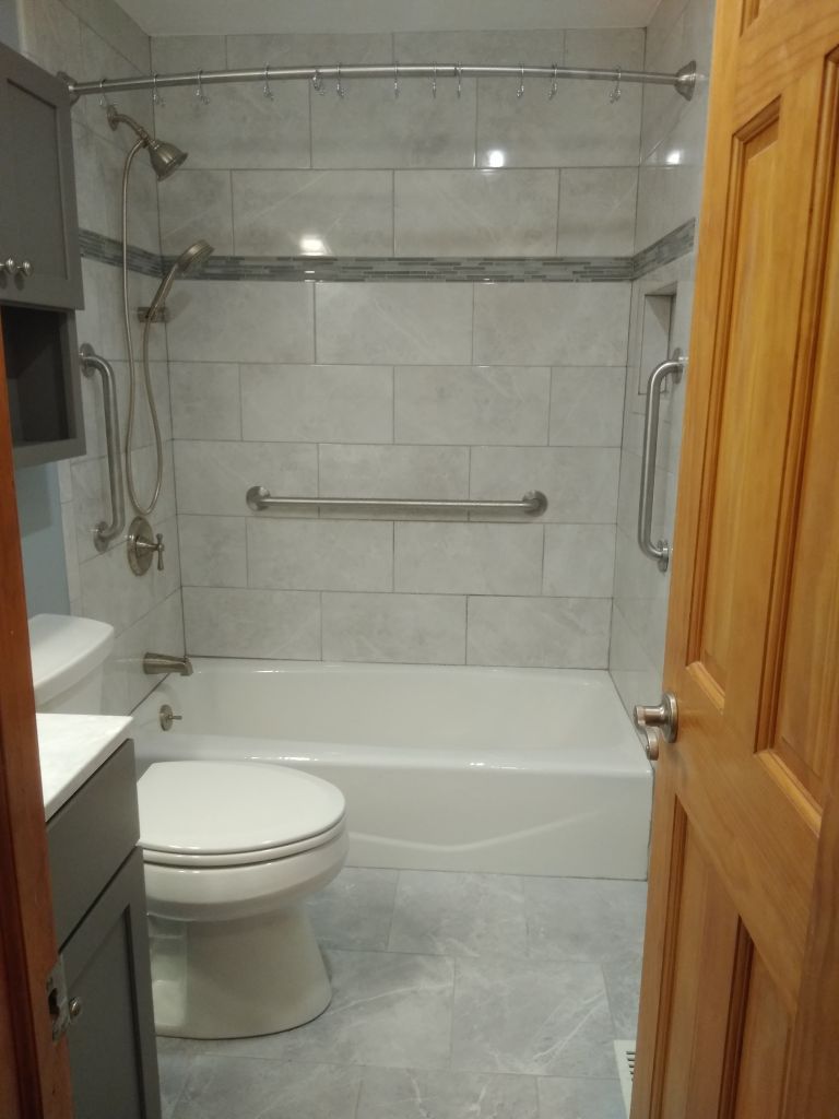 Small tiled bathroom with a white tub, toilet, grab bars, and glass shower doors on the left.