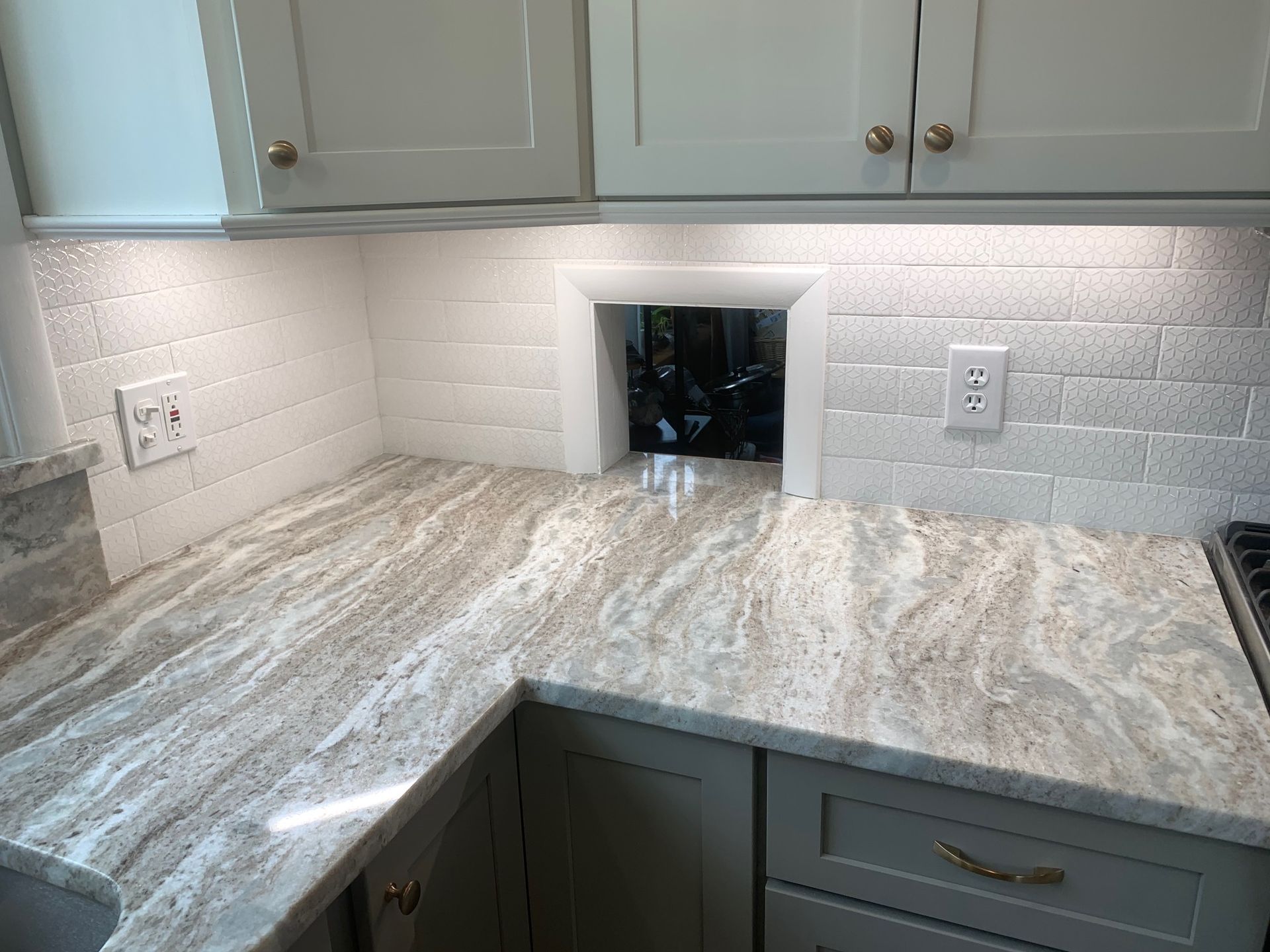 Marble-look kitchen countertop with gray cabinets, white subway tile backsplash, and a small wall opening.