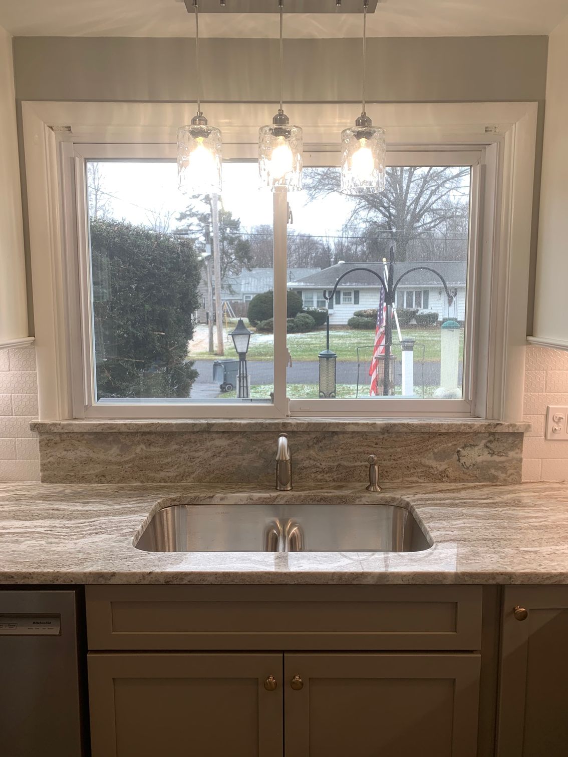 Kitchen sink beneath a window, with granite counters, chrome faucet, and pendant lights overlooking a street.