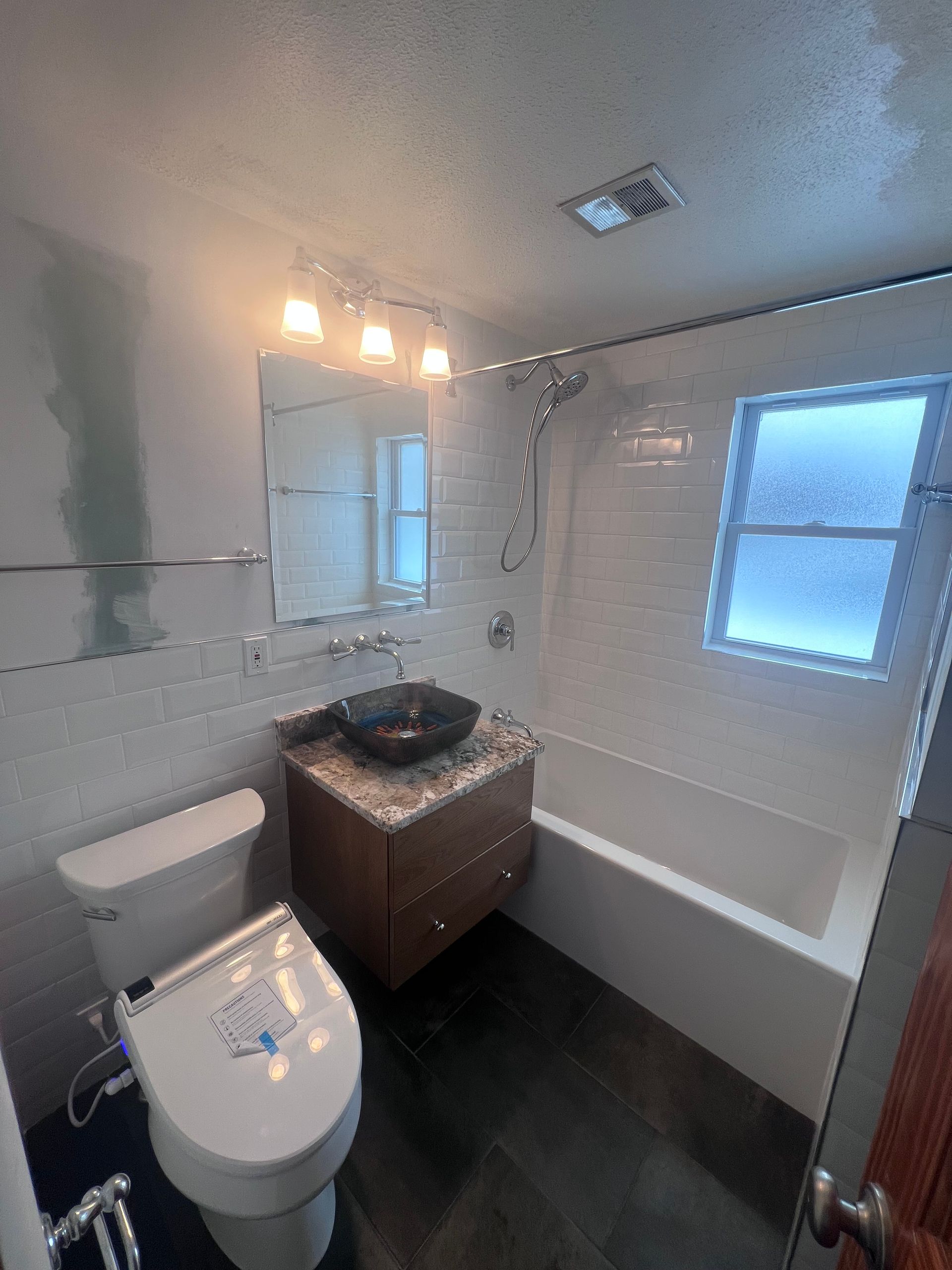 Small bathroom with toilet, vanity sink, bathtub, mirror lights, white walls, and dark floor.