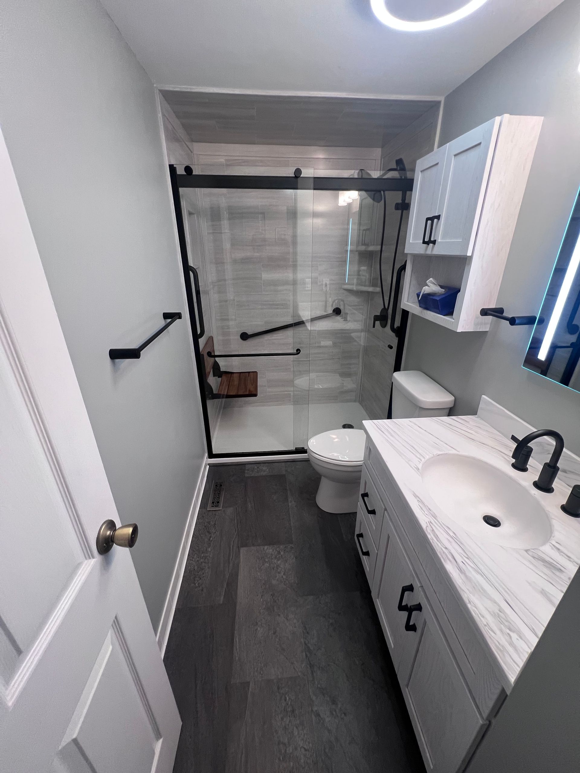 Narrow bathroom with white vanity, toilet, shower curtain, and dark floor runner under a ceiling ring light.