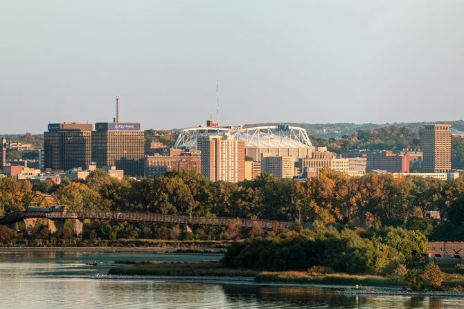 City skyline across a river with trees in the foreground and a stadium visible in the distance