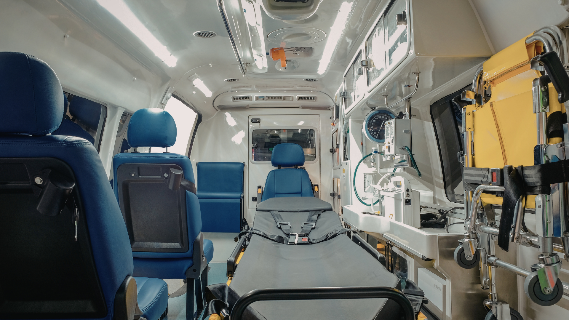 The interior of an ambulance featuring a central stretcher, blue passenger seats, medical equipment, and a yellow cot.