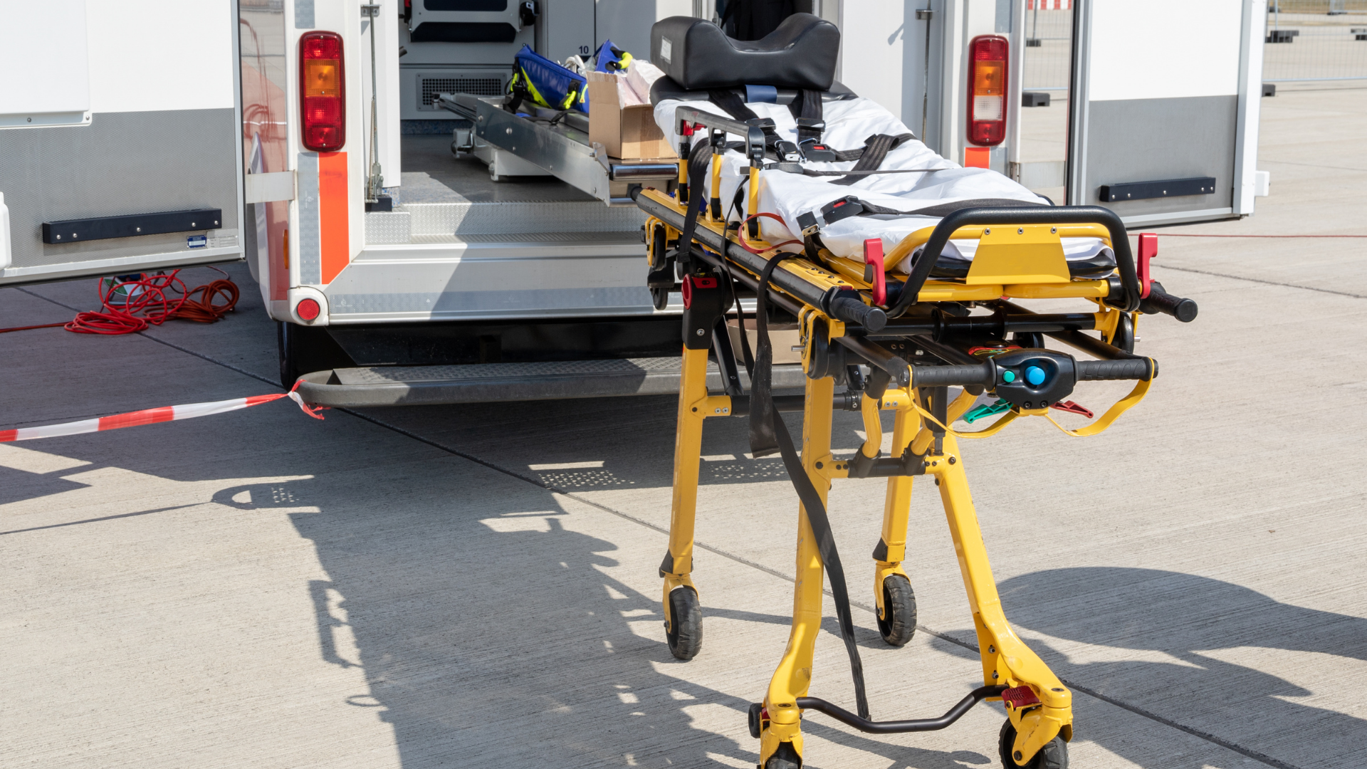 A yellow medical stretcher stands ready next to the open back doors of an ambulance parked on a concrete surface.