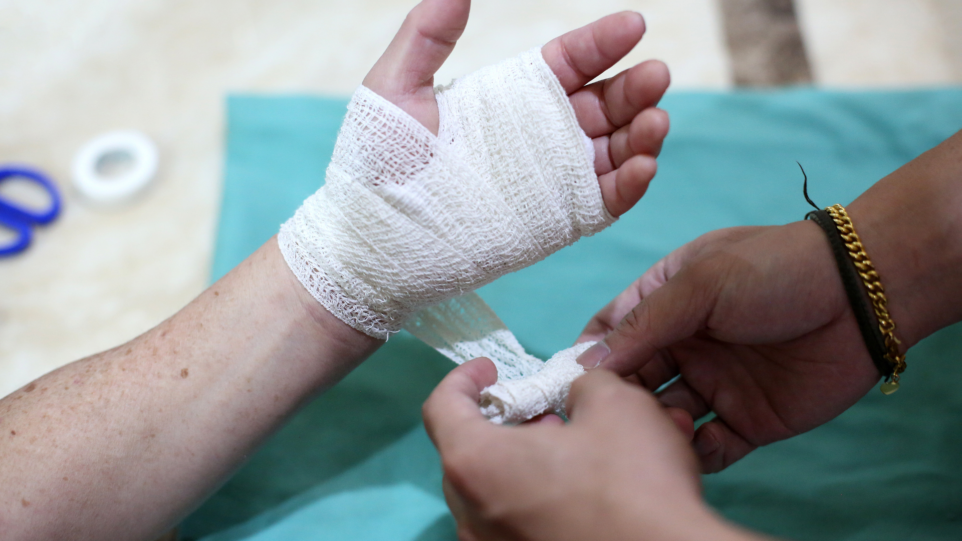 A person uses white gauze bandage to wrap a patient's hand and wrist on a green surface.