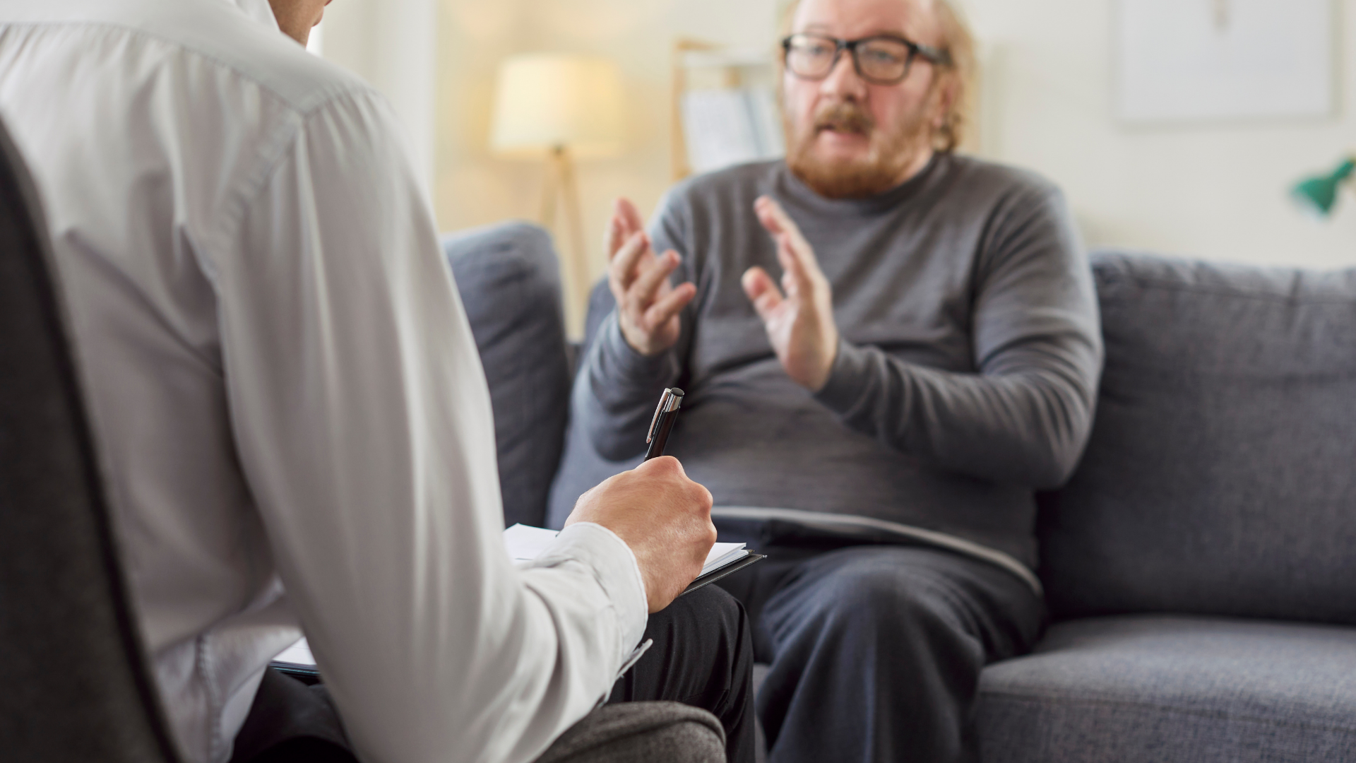 A professional in a white shirt takes notes while a person in a grey sweater gestures during a conversation on a sofa.