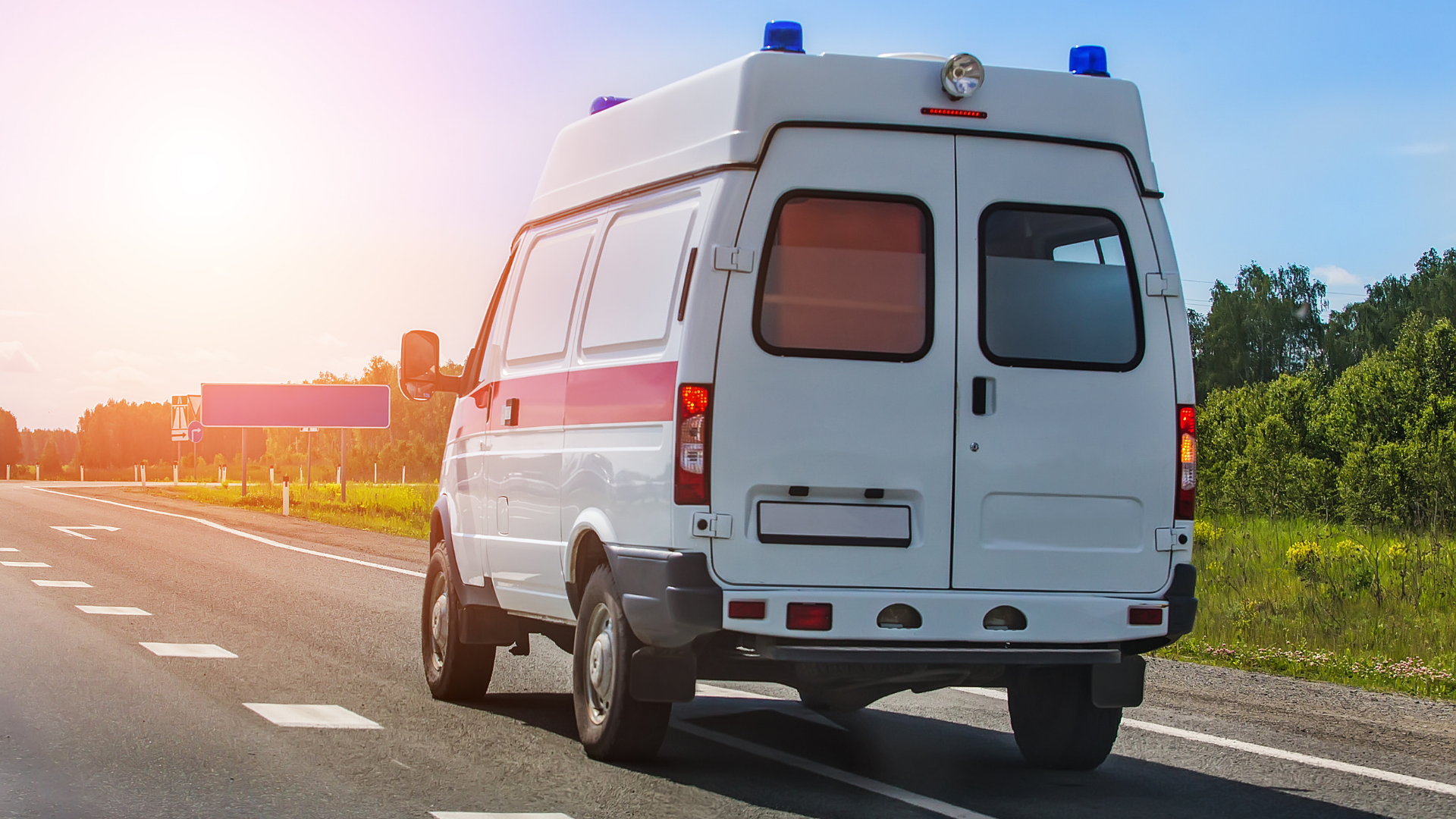 A white ambulance with red stripes drives along a rural road during a bright, sunny day.