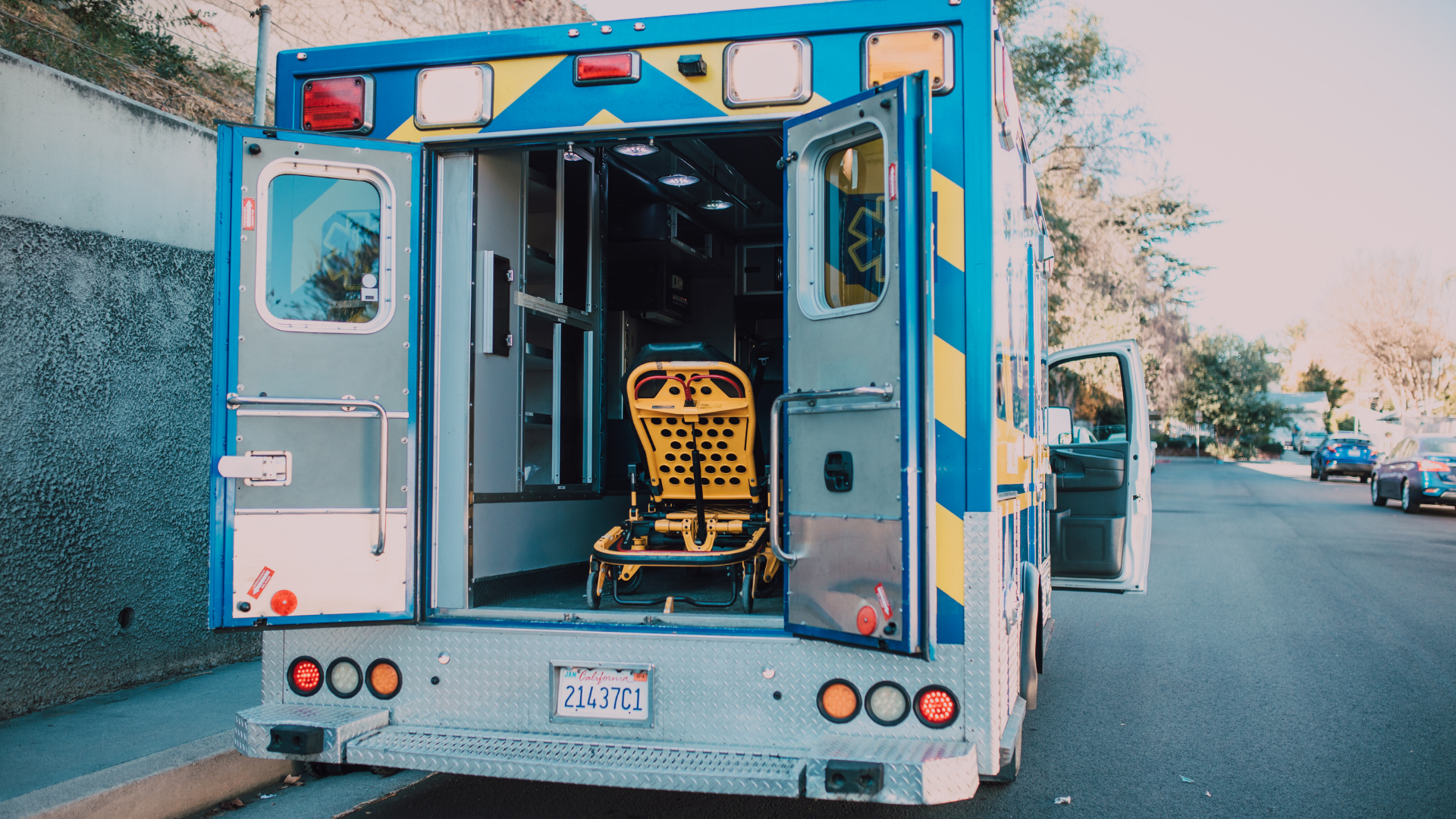 The back doors of a blue and white ambulance are open, showing a yellow stretcher parked on a street.