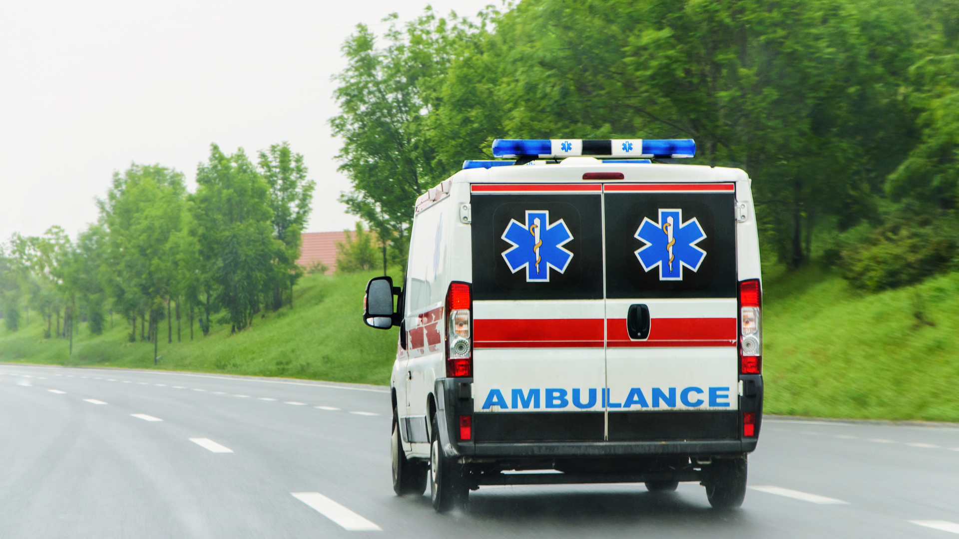 A white ambulance with red stripes and blue emergency lights travels down a road lined with green trees.