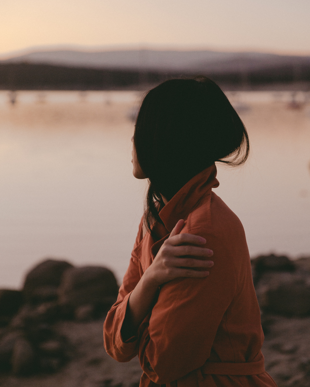 Person in a rust-colored coat stands by calm water at sunset, facing a blurred bridge in the distance