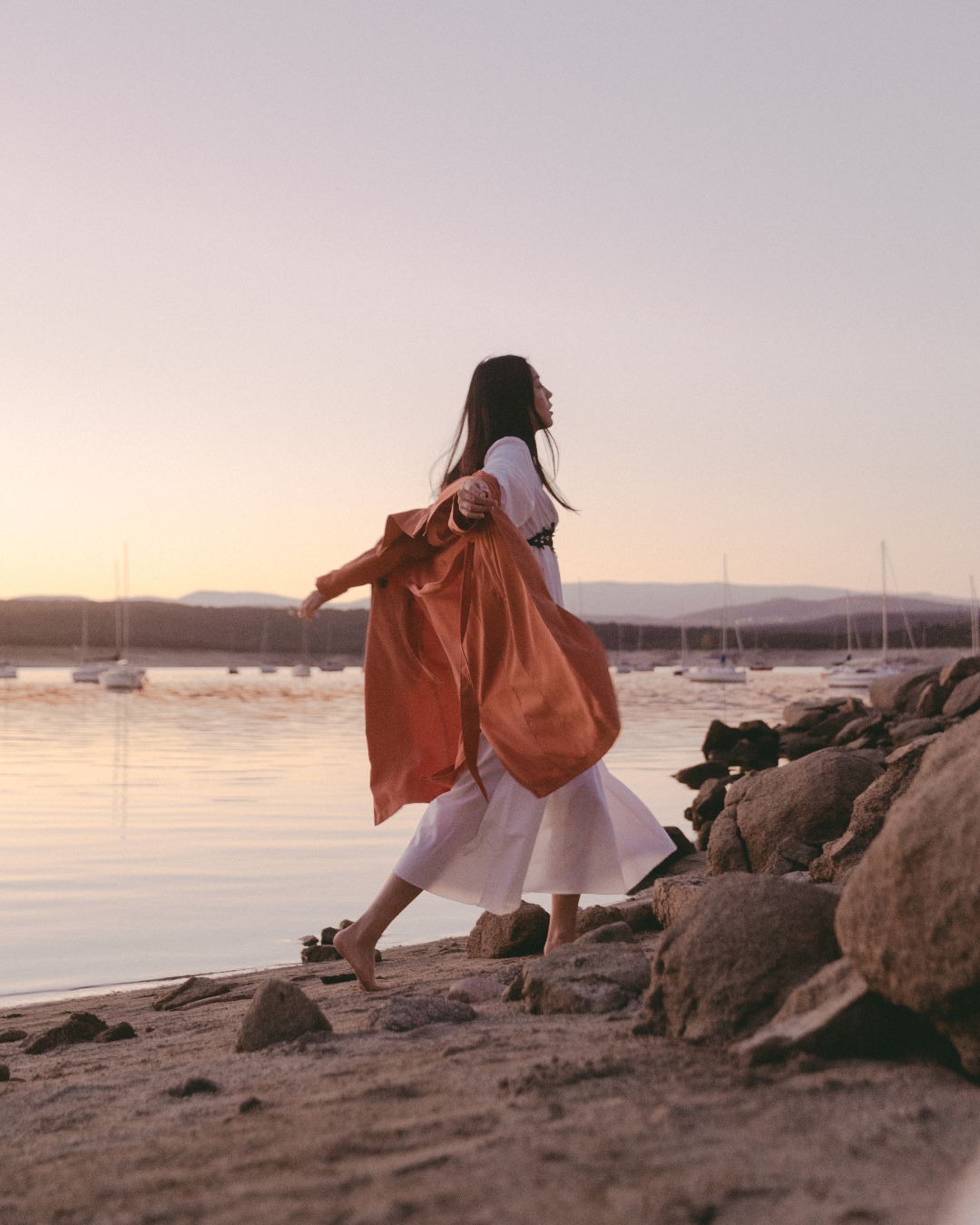 Person in a flowing white dress and orange shawl walking on a rocky beach at sunset.
