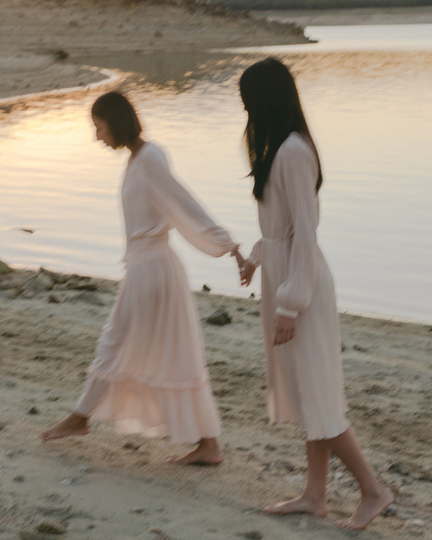 Two people in white dresses walking hand in hand along a beach at sunset