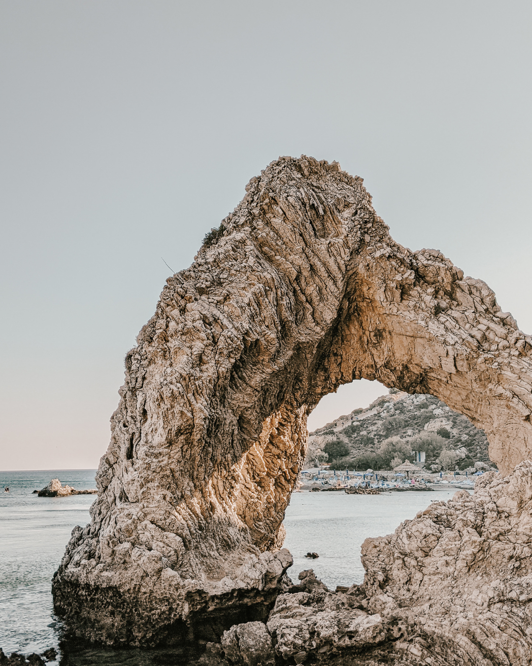 Rock arch rising from the sea against a pale sky, with distant boats and shoreline.