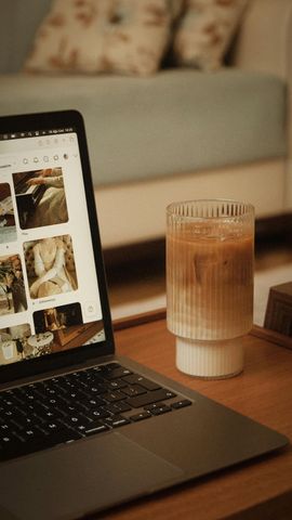 Laptop on a desk next to a tall iced drink, with a blurred sofa in the background.