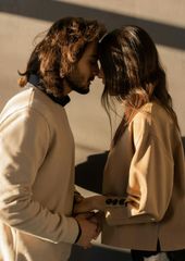 Two people in beige sweaters stand close, touching foreheads in warm sunlight.
