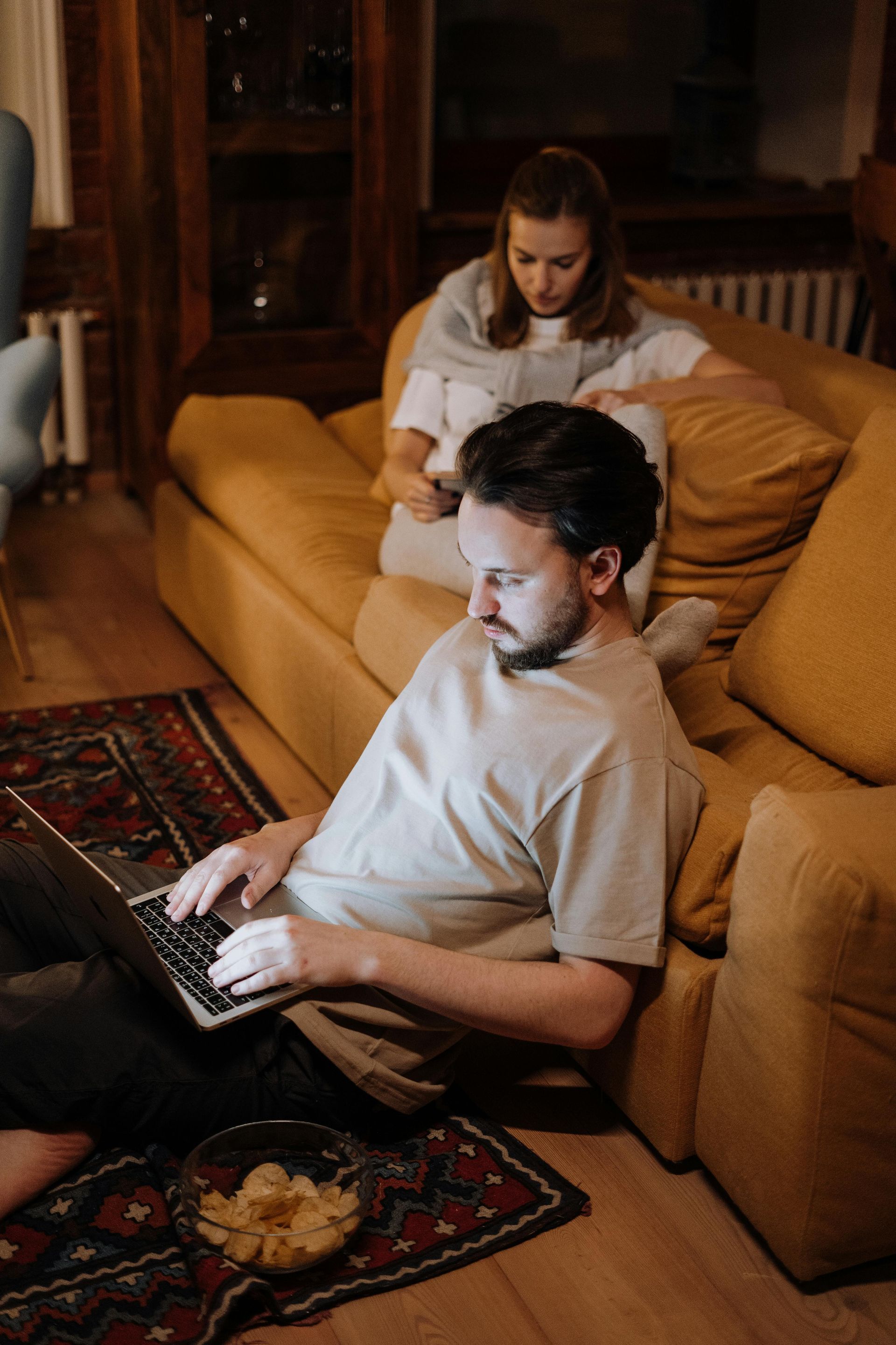 Two people on a mustard sofa, one using a laptop while the other sits behind them in a dim living room.