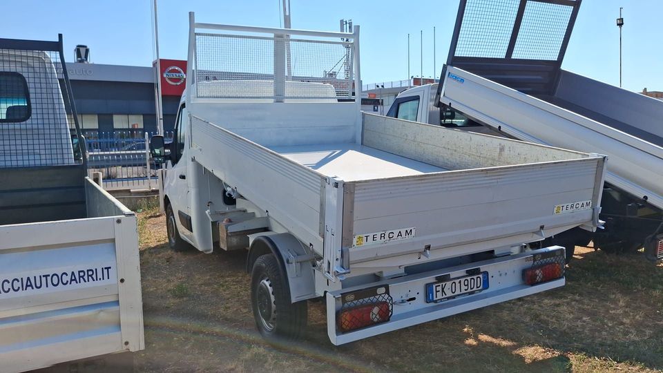 Un camioncino bianco con pianale ribassato, parcheggiato all'aperto accanto ad altri veicoli da lavoro in un piazzale soleggiato.