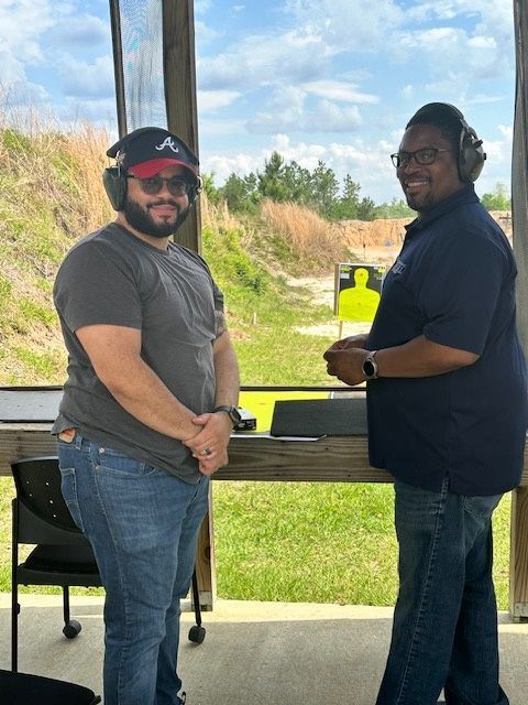 Two men at an outdoor shooting range. One wears a cap, the other a blue shirt. Both have ear protection and are smiling.