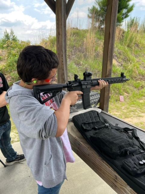 Teen shooting rifle at outdoor range, wearing ear and eye protection. Rifle case nearby.