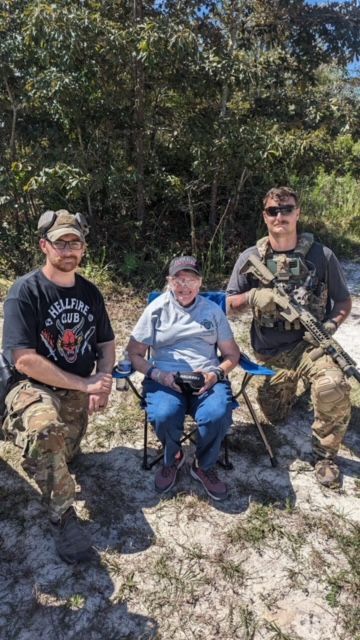 Three people, one seated, pose outdoors: two men, one wearing camouflage, a seated person in a chair. Sunny day.
