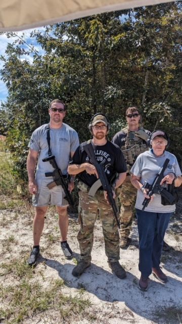 Four people at an outdoor shooting range hold rifles. One wears camouflage, others shorts and casual clothes.
