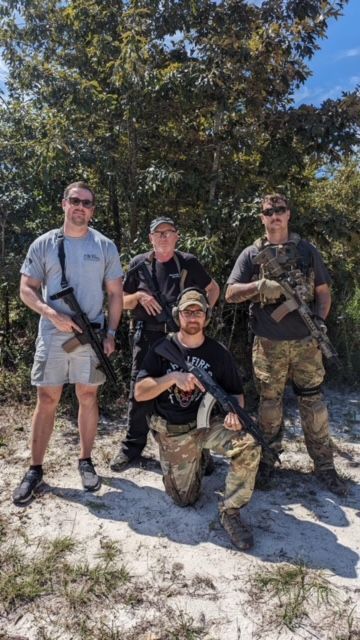 Four men with rifles pose outdoors; one kneels. Sunny, trees in background.