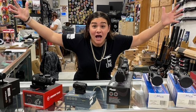 Woman in gun store excitedly displays scopes with arms outstretched.