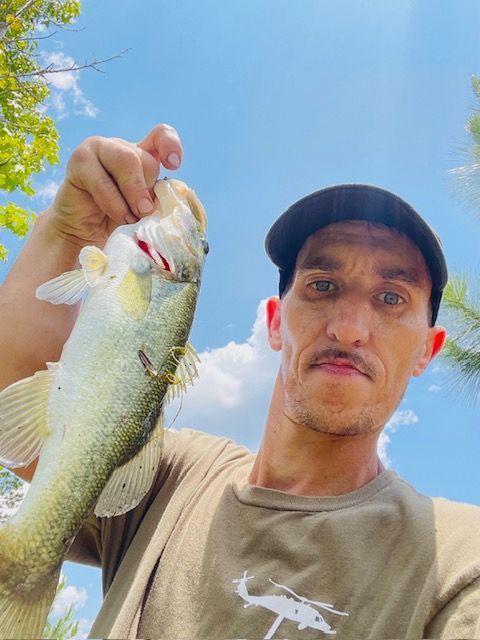 Man holding a bass fish under a blue sky. He wears a hat and a tan shirt, smiling.