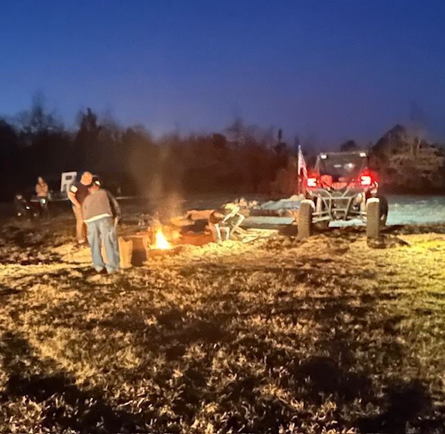 People gathered around a bonfire in a field next to an off-road vehicle at dusk.