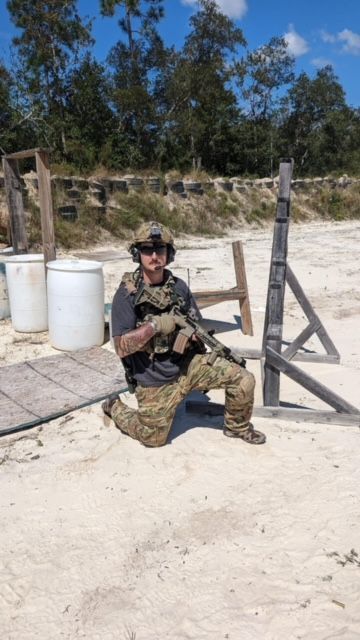 Man kneeling on a shooting range, holding a rifle. Wearing camouflage, helmet, and body armor.
