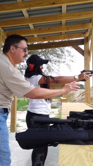 Man assisting a woman at an outdoor shooting range, both holding guns, woman aiming.