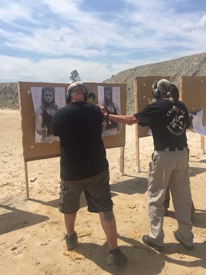 Two men at a shooting range; one aiming at a target, the other assisting, with posters of a person on the targets.