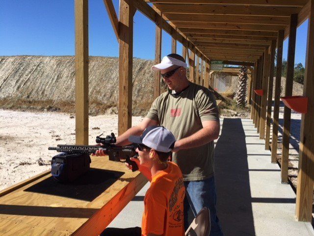 Man teaching a boy to shoot a rifle at an outdoor shooting range.