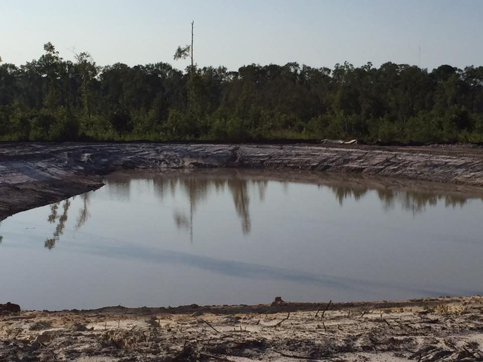 Pond with still water reflects trees and sky, surrounded by dirt banks.